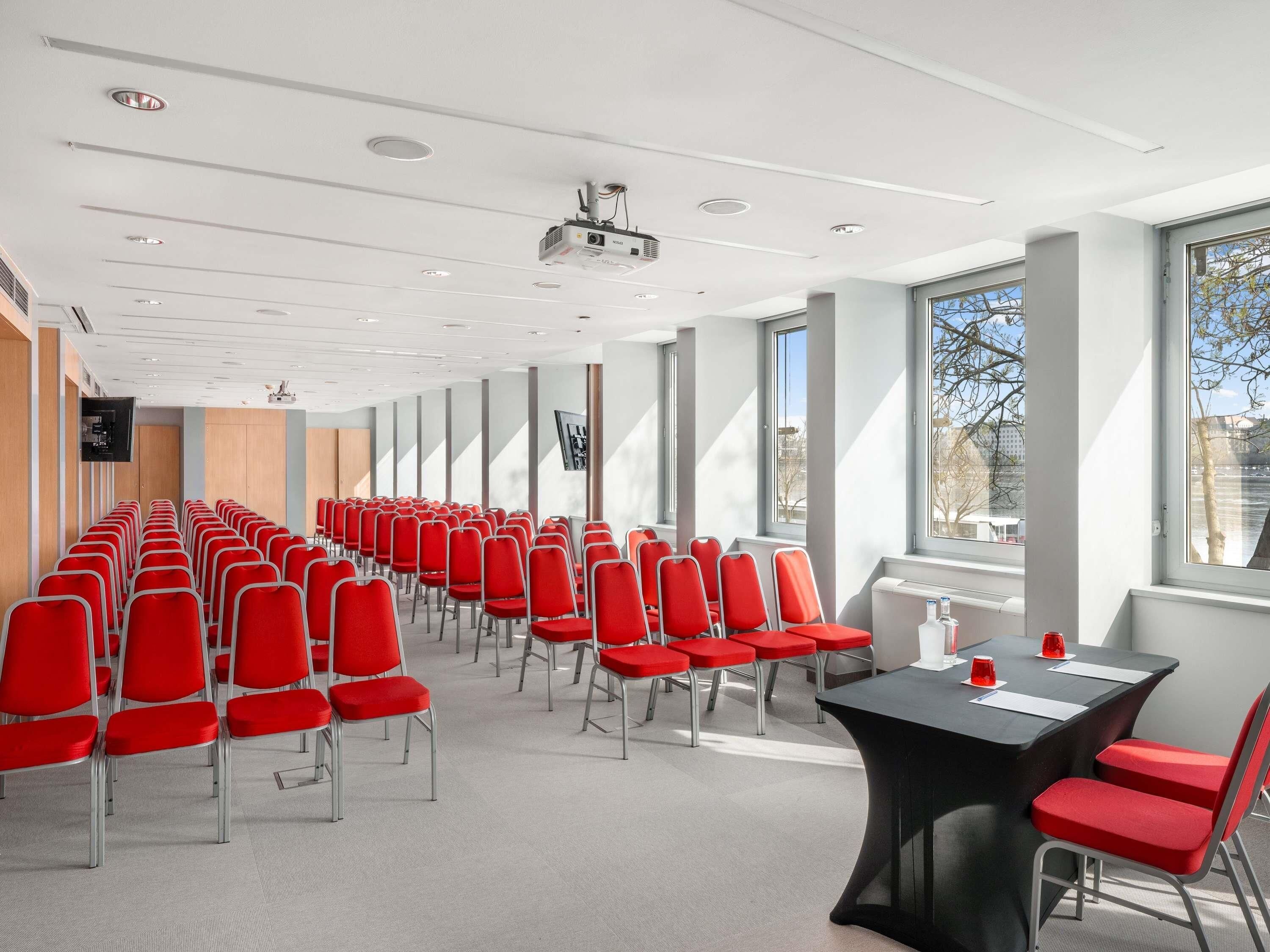 Ein Konferenzraum mit Reihen roter Stühle, einem Projektor und großen Fenstern.