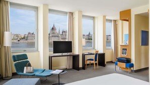 A hotel room with a large bed, a desk with a computer, a chair, and a view of a historic building through the windows. - Novotel Budapest Danube (Budapest)