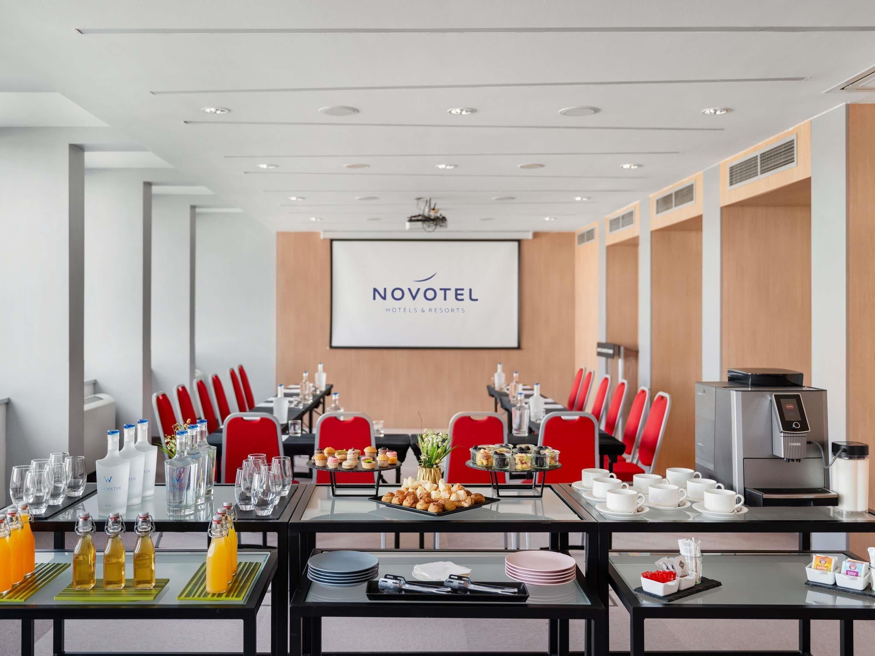 A conference room with a long table, red chairs, and a projector screen displaying 'NOVOTEL HOTELS & RESORTS'.