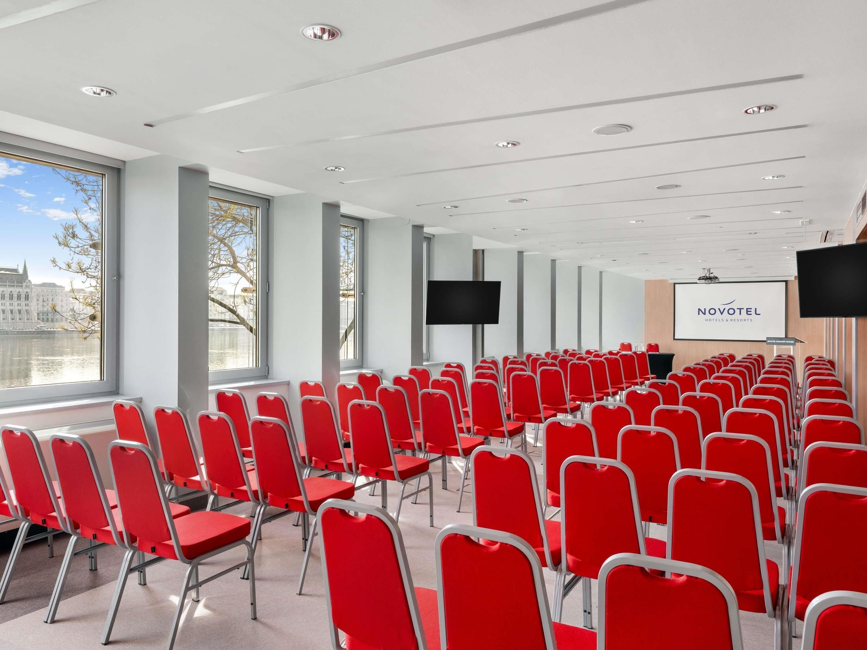 A conference room with rows of red chairs, large windows, a projector screen, and a Novotel sign.