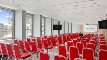 A conference room with rows of red chairs, large windows, a projector screen, and a Novotel sign.