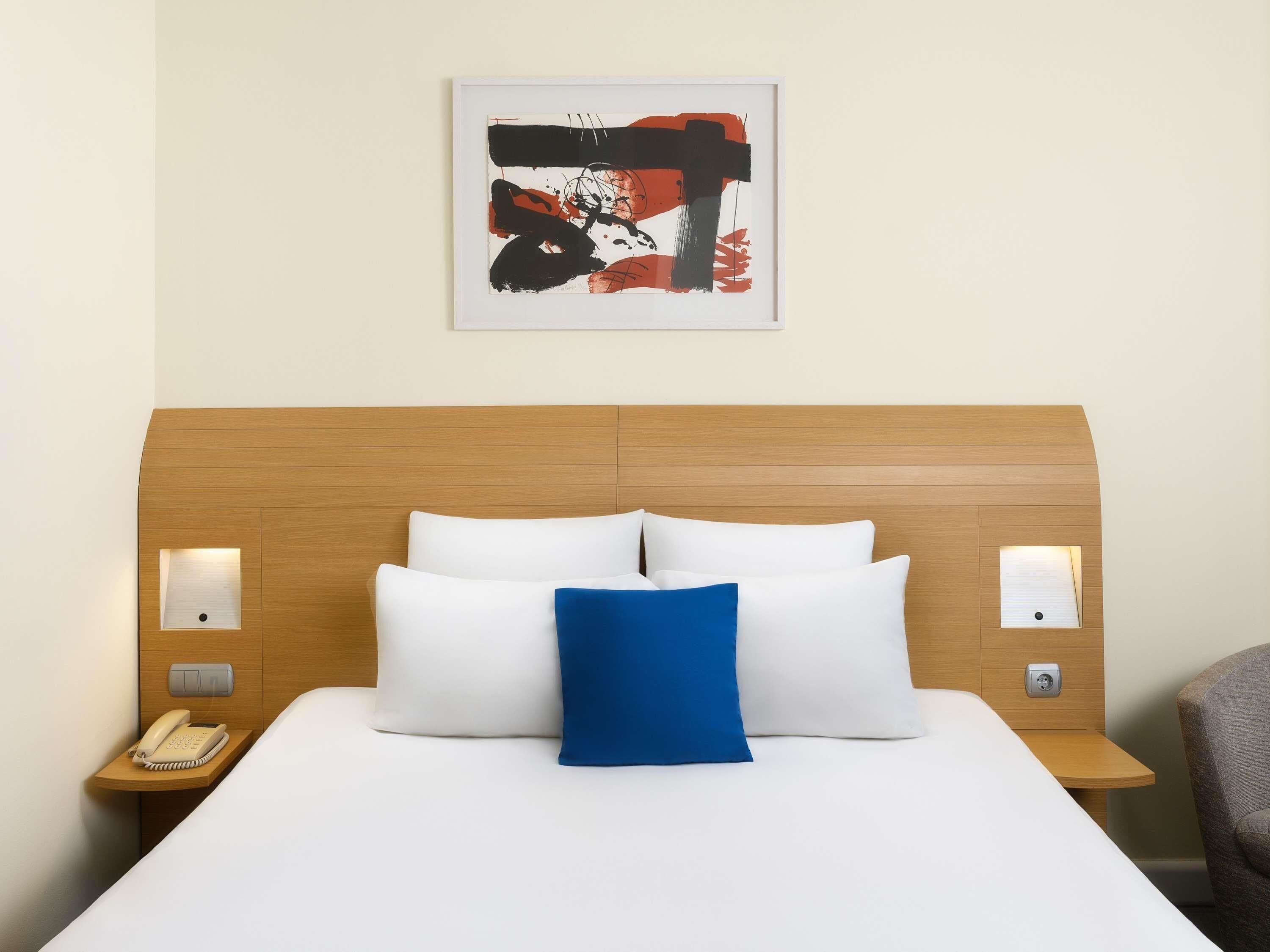A hotel room with a wooden headboard, a bed with white and blue pillows, a bedside table with a phone, and a wall-mounted artwork.