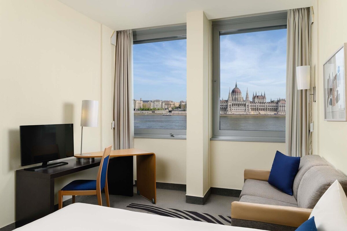 A hotel room with a bed, a desk with a chair, a television, and large windows offering views of a historic building and a river.
