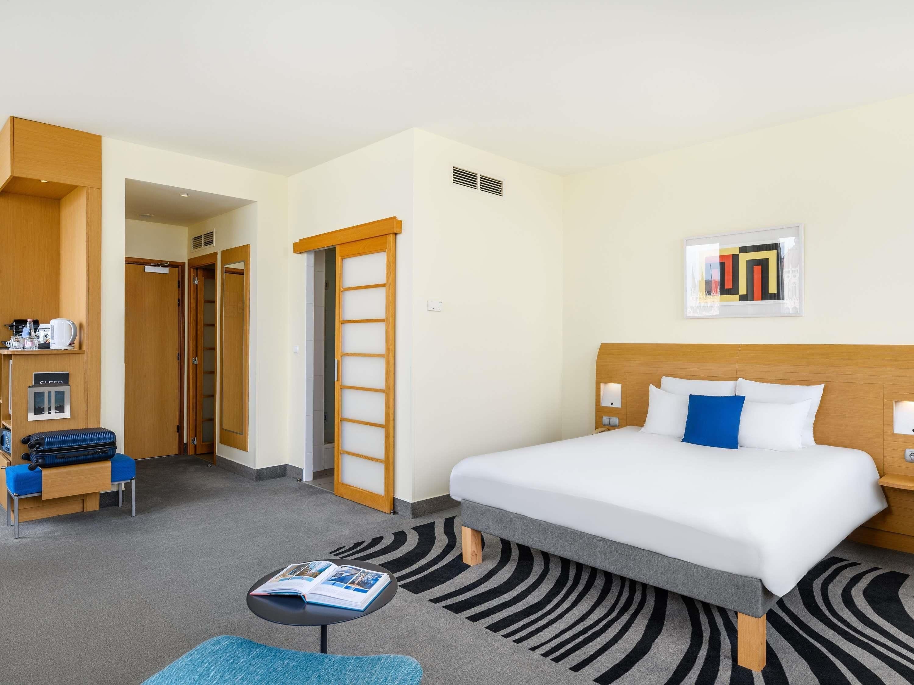 A hotel room with a bed, a desk, a chair, a coffee machine, and a bookshelf.