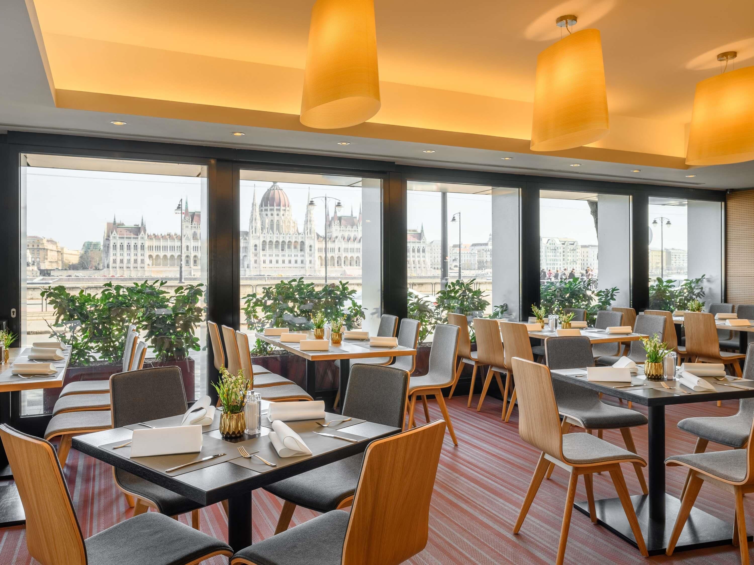 Un restaurante con mesas y sillas, ventanas amplias y plantas en macetas.