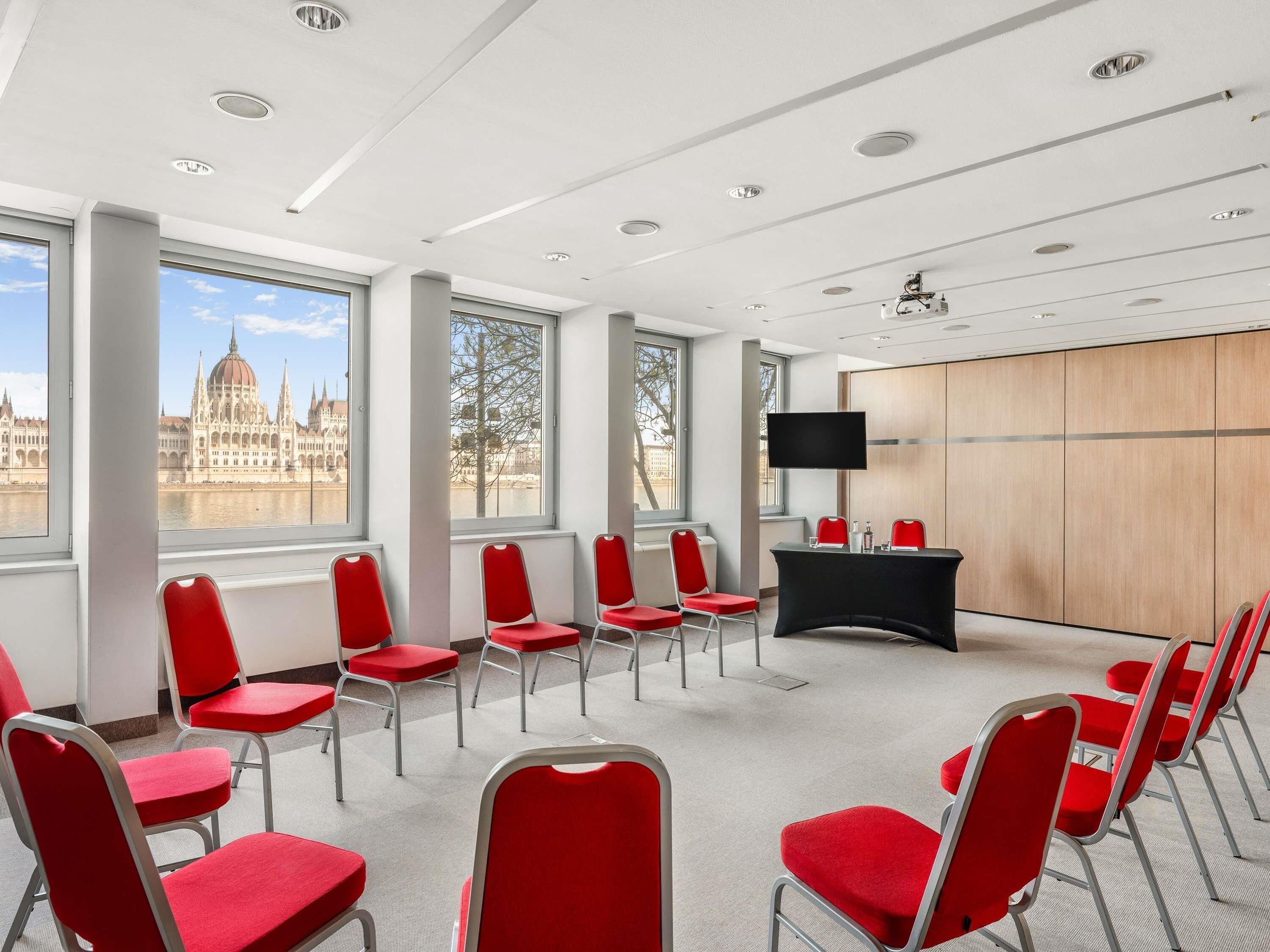 Una sala de conferencias con sillas rojas, una mesa negra y un amplio ventanal con vistas a un edificio histórico.