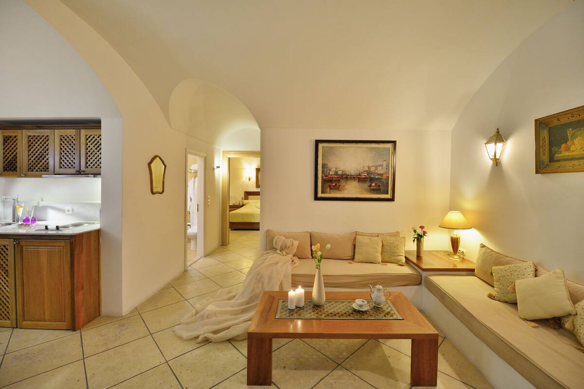 A living room with a sofa, a coffee table with candles, a painting on the wall, and a kitchen area with cabinets and a sink.