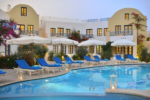 Un área de piscina con sillones de descanso y sombrillas, una piscina cristalina y un edificio con balcones y ventanas.