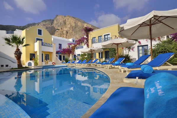 Un área de piscina con sillas reclinables azules y sombrillas, una piscina azul cristalina y una montaña al fondo.