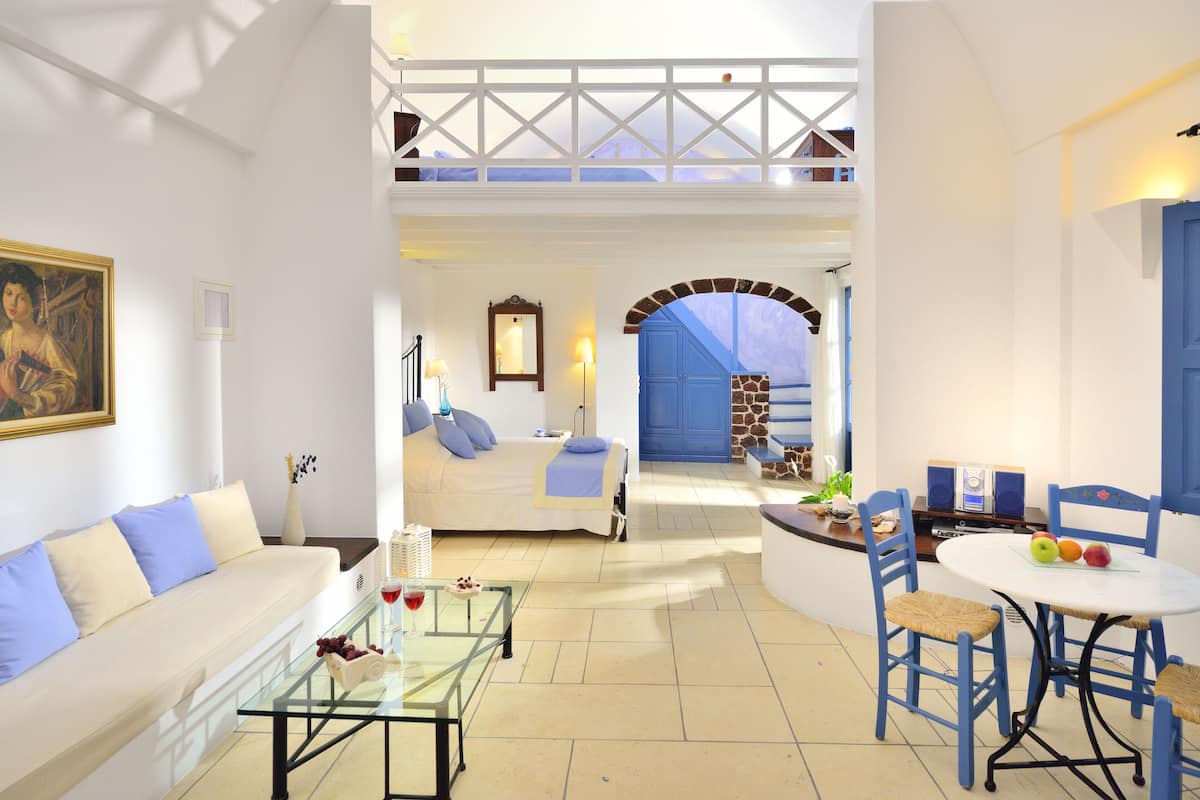 A spacious living area with a sofa, coffee table, and dining table with chairs. There is a bed with a view to the outside, a blue door, and a staircase leading to an upper level.