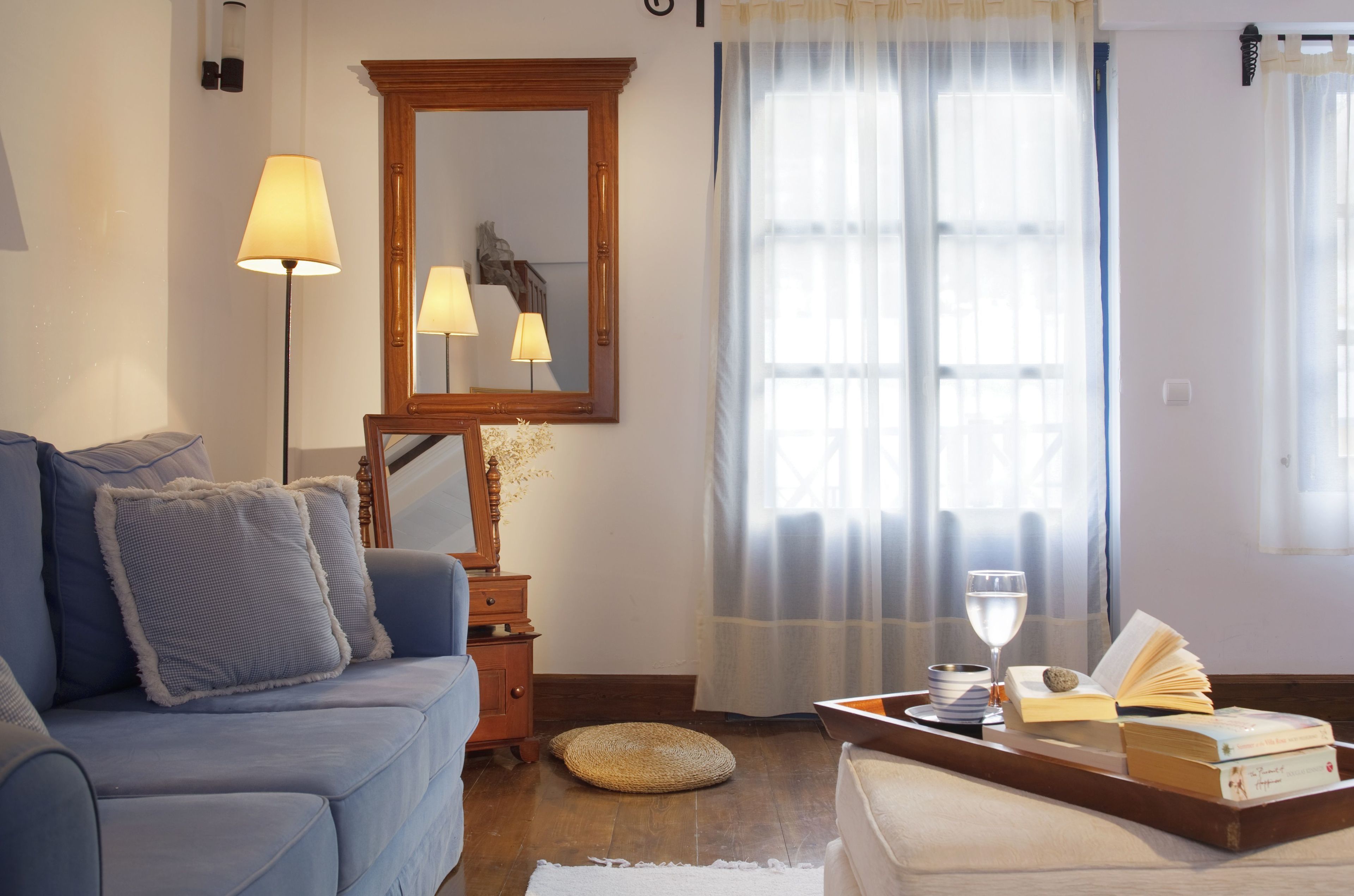 A living room with a blue sofa, a wooden side table with a lamp, a mirror, and a window with sheer curtains.