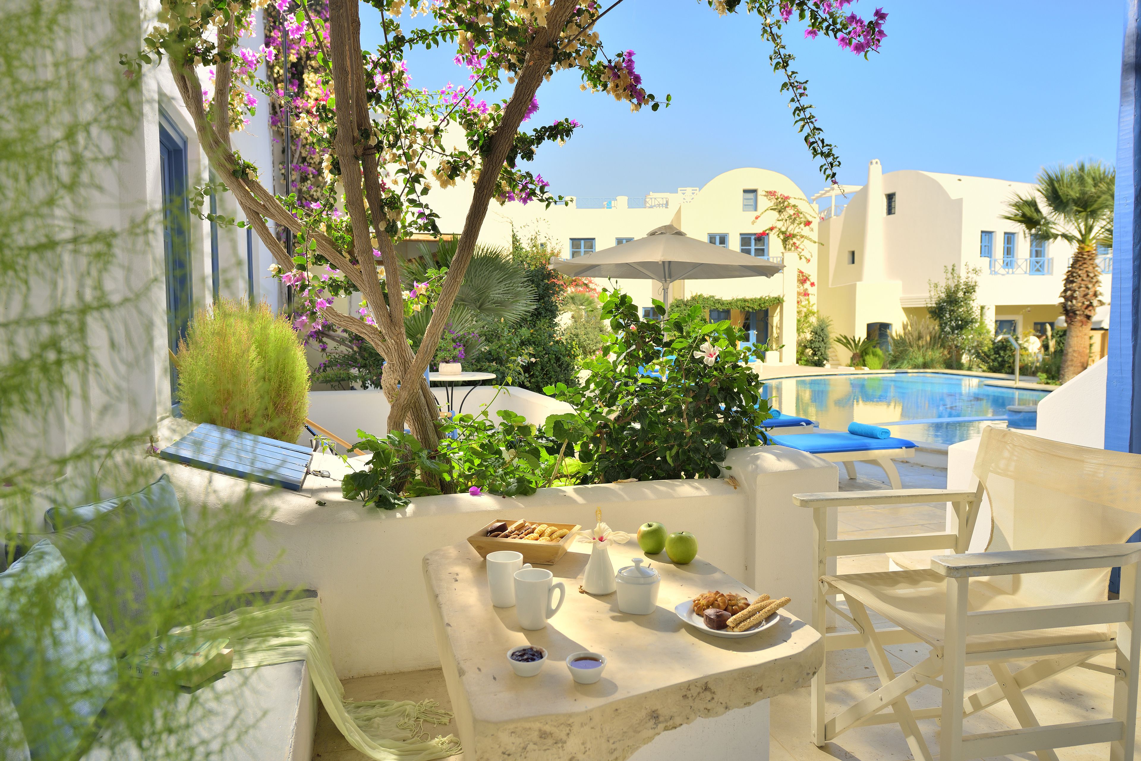 Una terraza con una mesa preparada para el desayuno, una piscina y edificios blancos al fondo.
