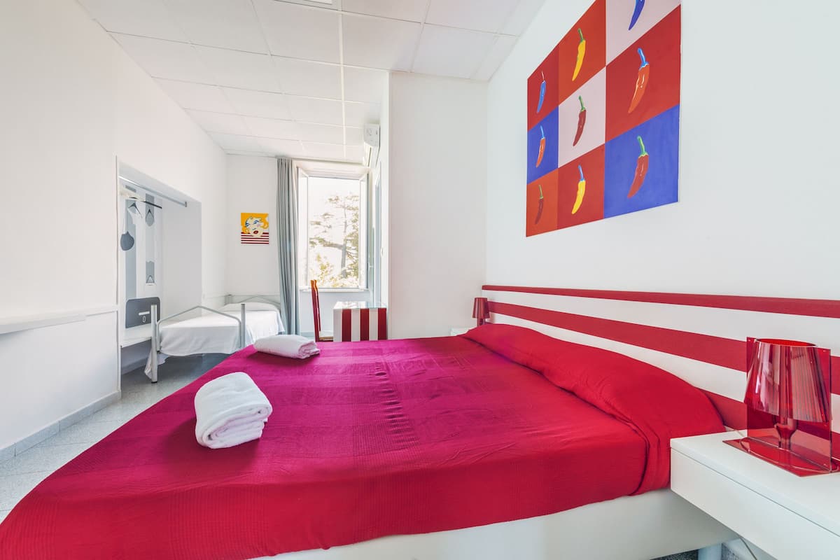 A hotel room with a red bedspread, a white bedside table with a red lamp, and a wall art of chili peppers.