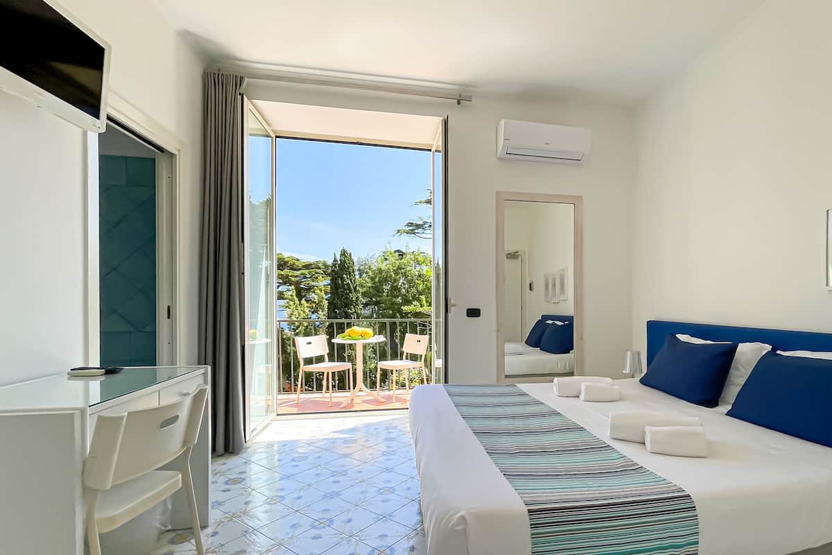 A hotel room with a bed, a desk, a chair, a balcony with a table and chairs, and a view of trees and sky.