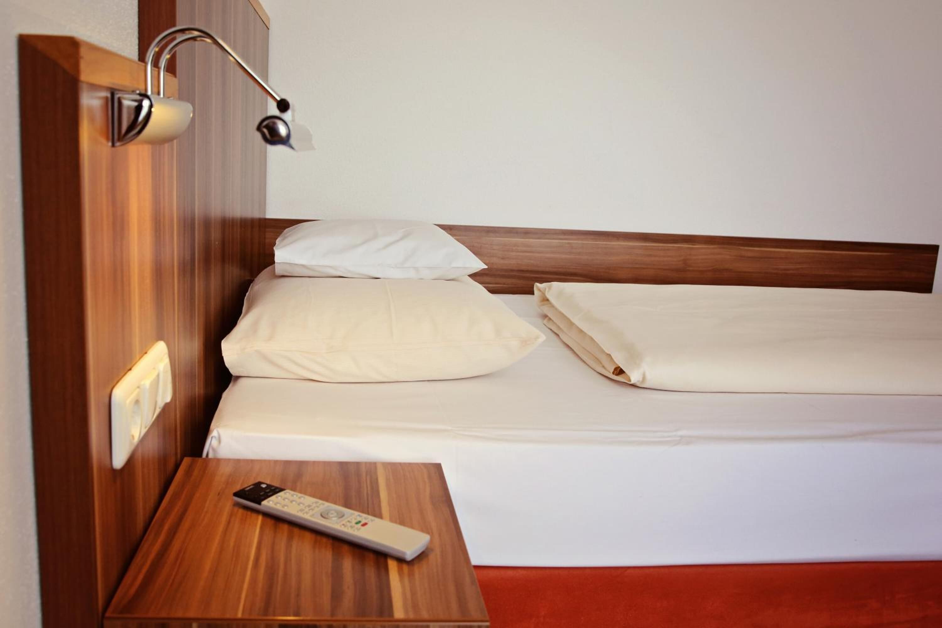 A neatly made bed with white linens, a wooden nightstand with a remote control, and a wall-mounted lamp.