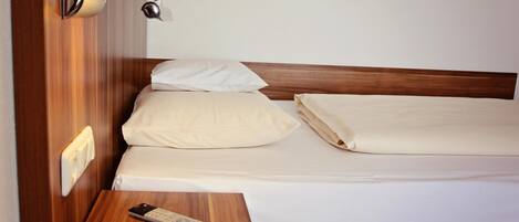 A neatly made bed with white linens, a wooden nightstand with a remote control, and a wall-mounted lamp.