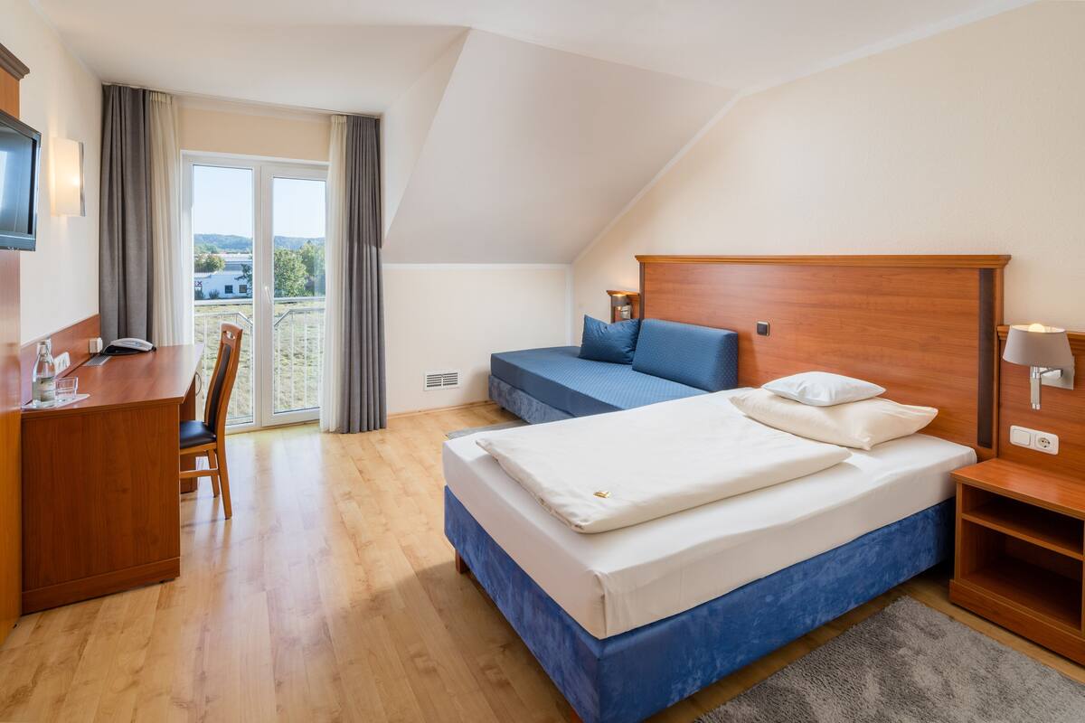 A bedroom with a bed, a desk, and a chair. There is a balcony with curtains.