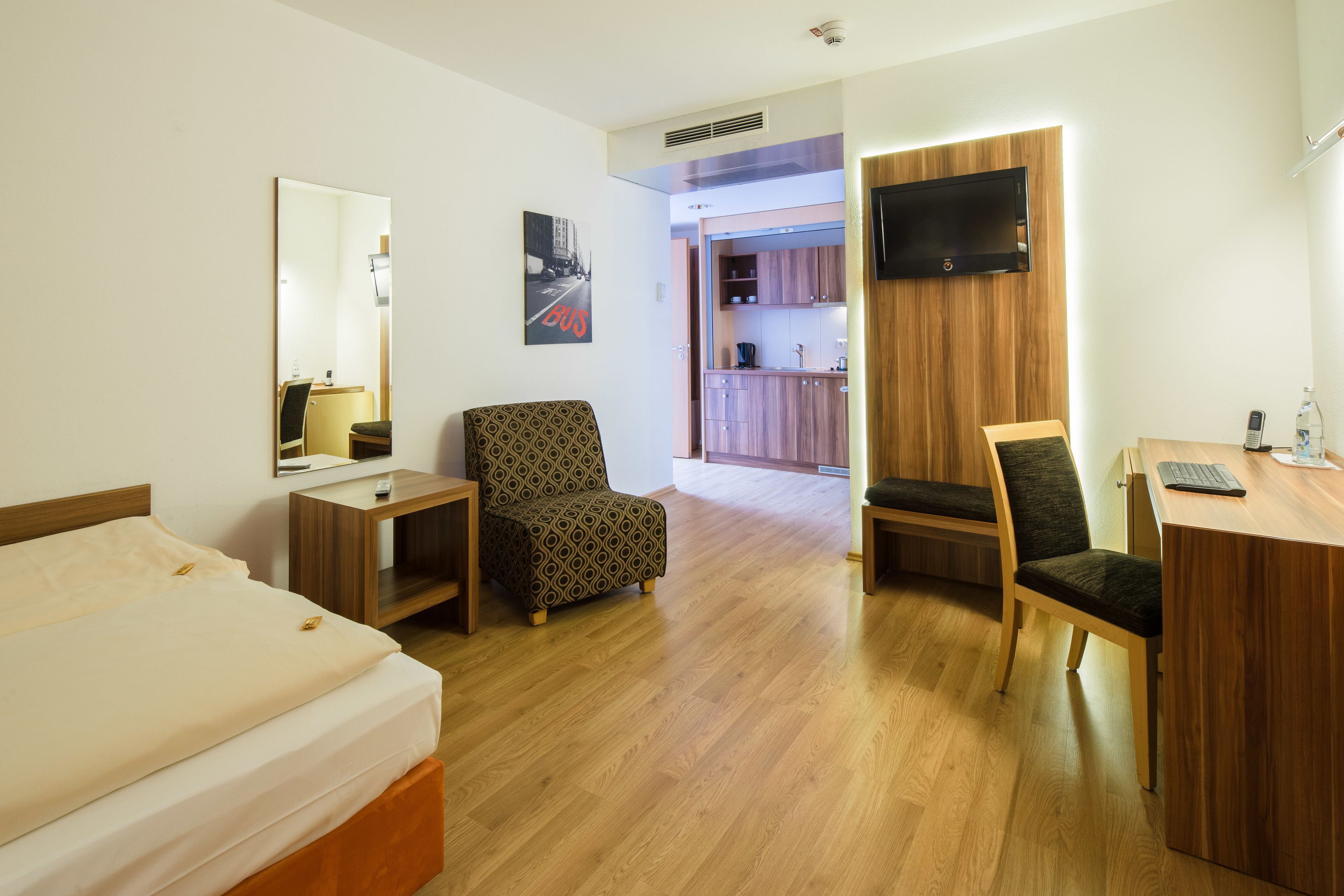 Habitación de hotel con una cama, dos sillas, un escritorio, un televisor y una kitchenette al fondo.