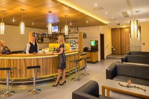 Uma área de bar com balcão de madeira, banquetas e um espaço de estar com sofá e mesas.
