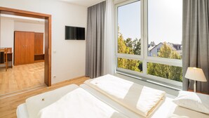 A hotel room with a bed, a TV mounted on the wall, a window with curtains, and a view of trees and buildings outside. - Hotel Meridian - Landshut (Ergolding)