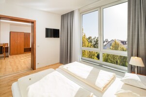 Quarto de hotel com cama, TV fixada na parede, janela com cortinas e vista para árvores e edifícios.
