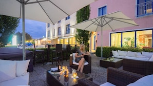 An outdoor seating area with wicker furniture and umbrellas, a woman sitting on a chair, and a building with illuminated windows in the background. - Hotel Meridian - Landshut (Ergolding)