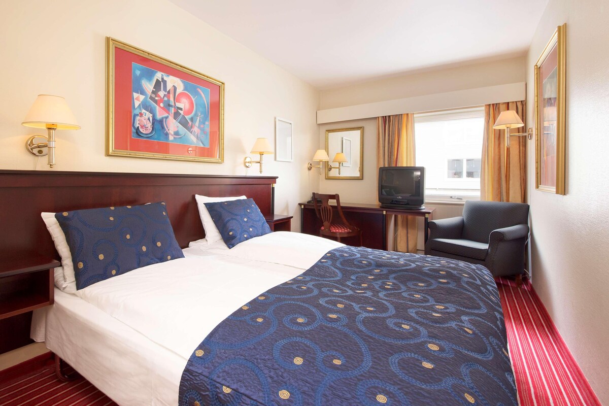 A hotel room with a large bed, a desk with a television, a chair, and a window with curtains.