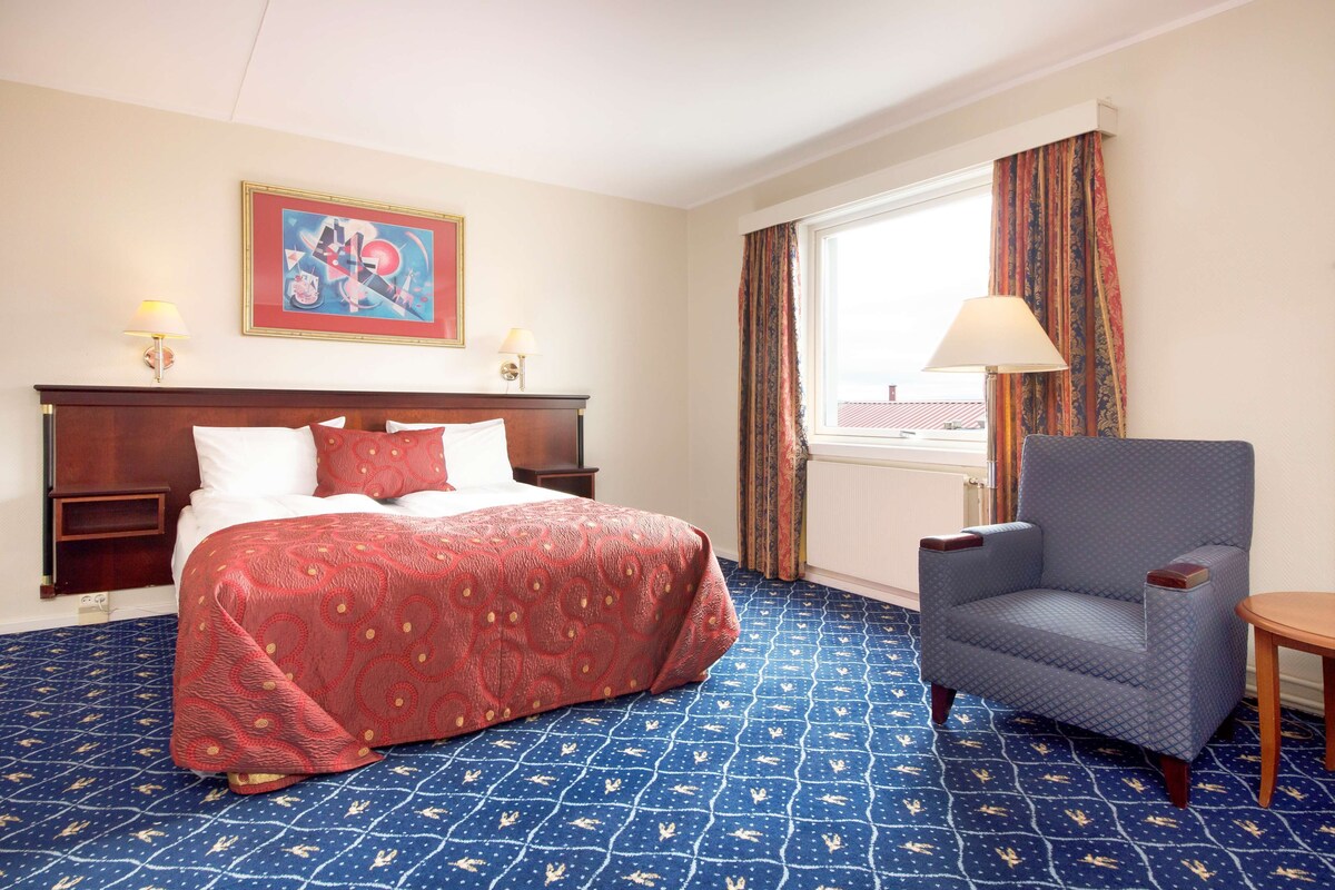 Quarto de hotel com cama, uma cadeira, luminária, quadro e janela com cortinas.
