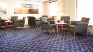Una habitación de hotel con una alfombra azul estampada, varios sillones, una mesita con una lámpara y un cuadro grande de un atardecer.