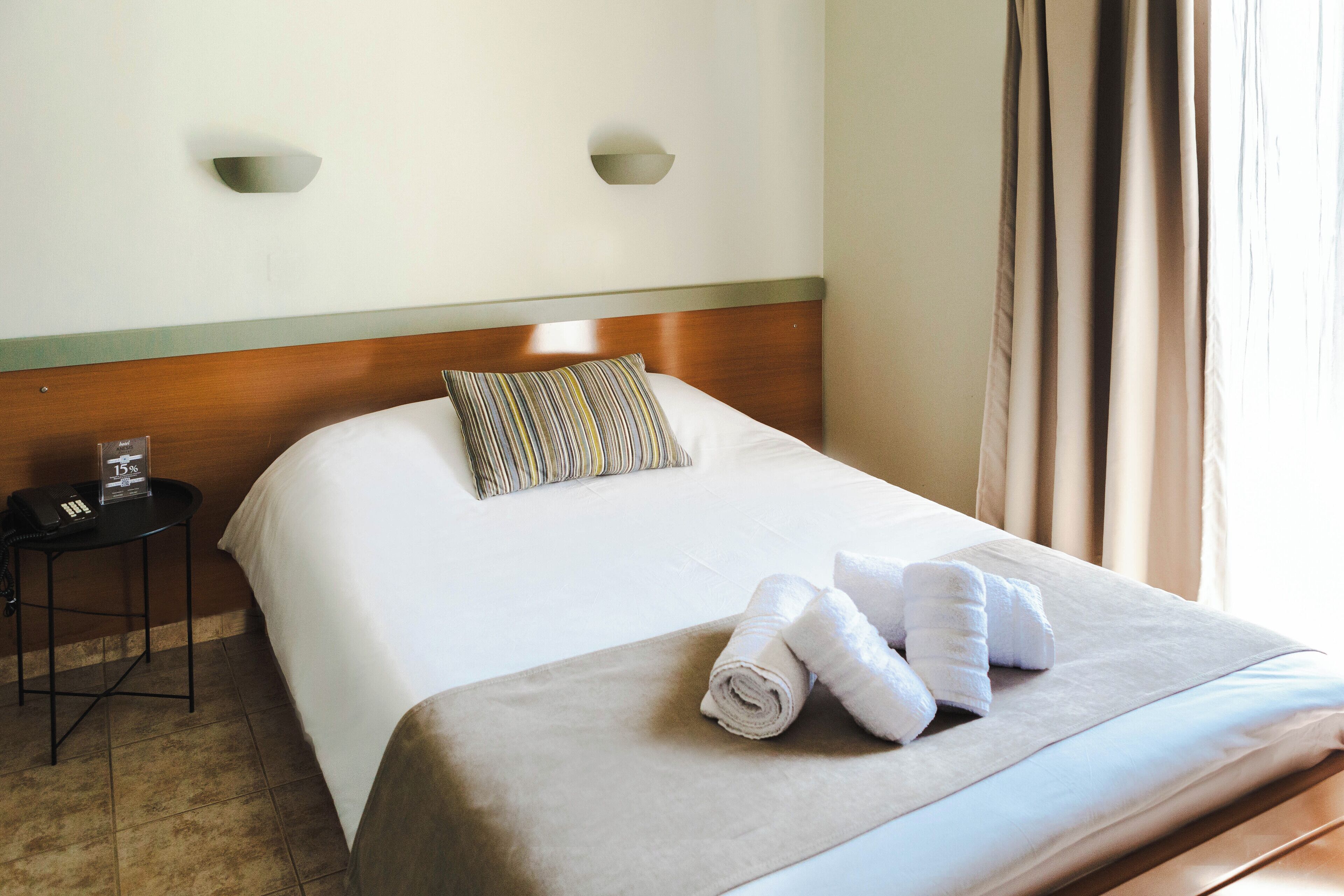 Una cama bien hecha con un cojín a rayas, una toalla enrollada y una mesita de noche con un teléfono y un vaso.