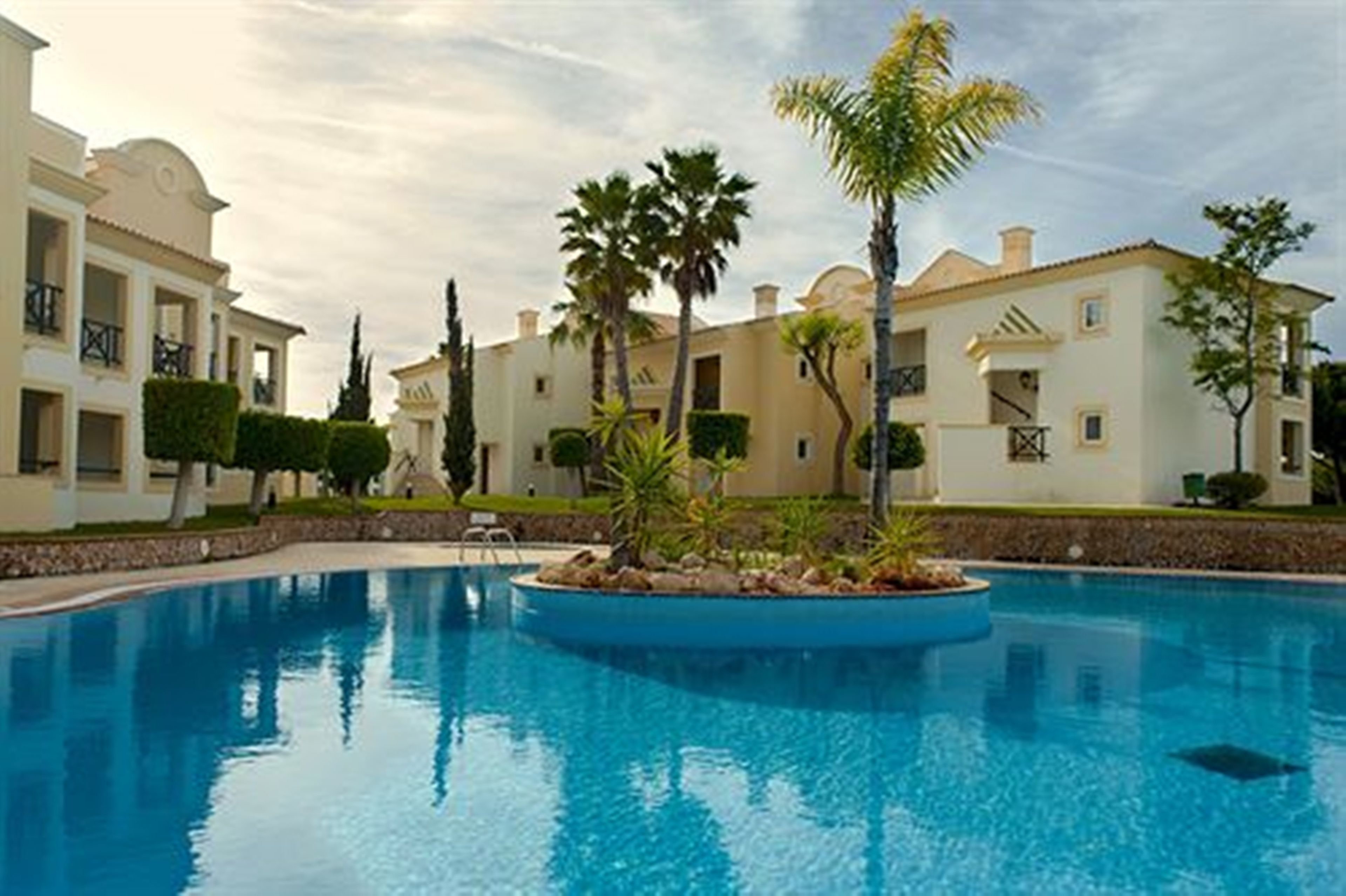 A swimming pool with a circular island, surrounded by palm trees and a building with a Mediterranean architectural style.