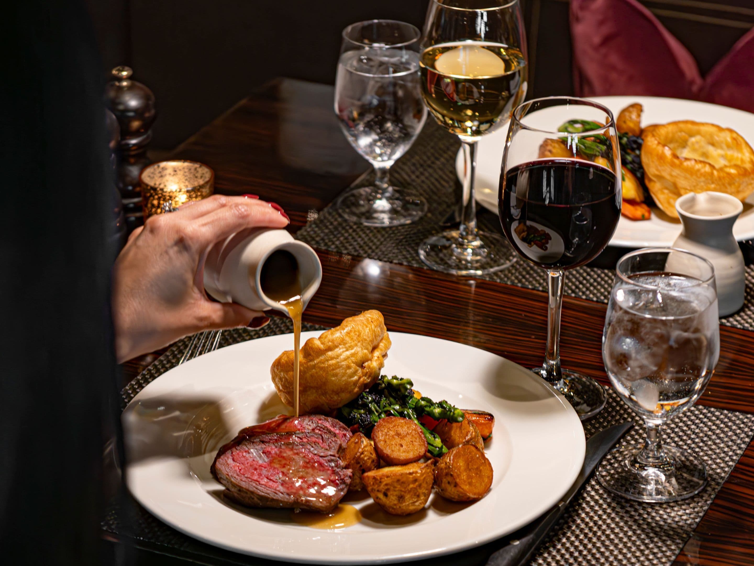 Una mesa de comedor con un plato de comida, una copa de vino y una persona sirviendo una bebida.