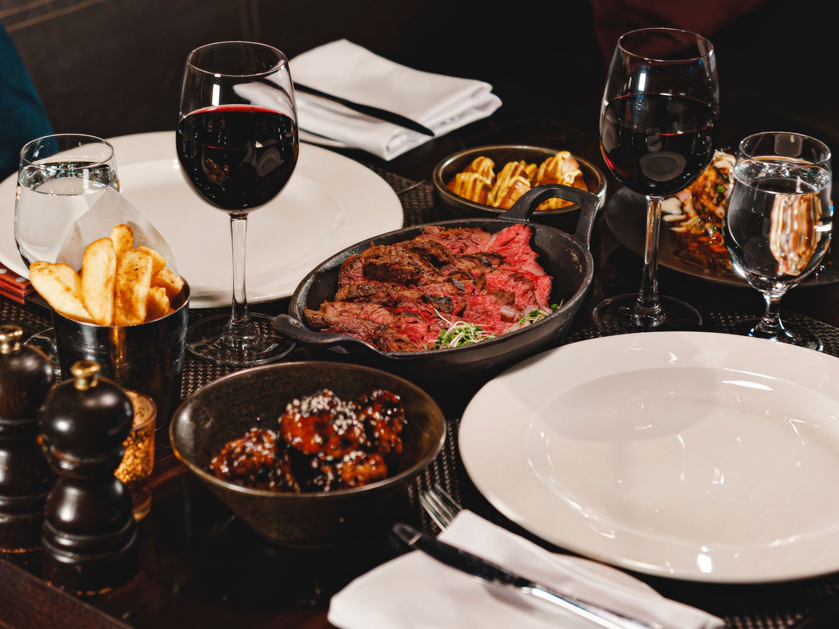 A dining table with various dishes, including steak, fries, and a dessert, along with multiple glasses of red wine.