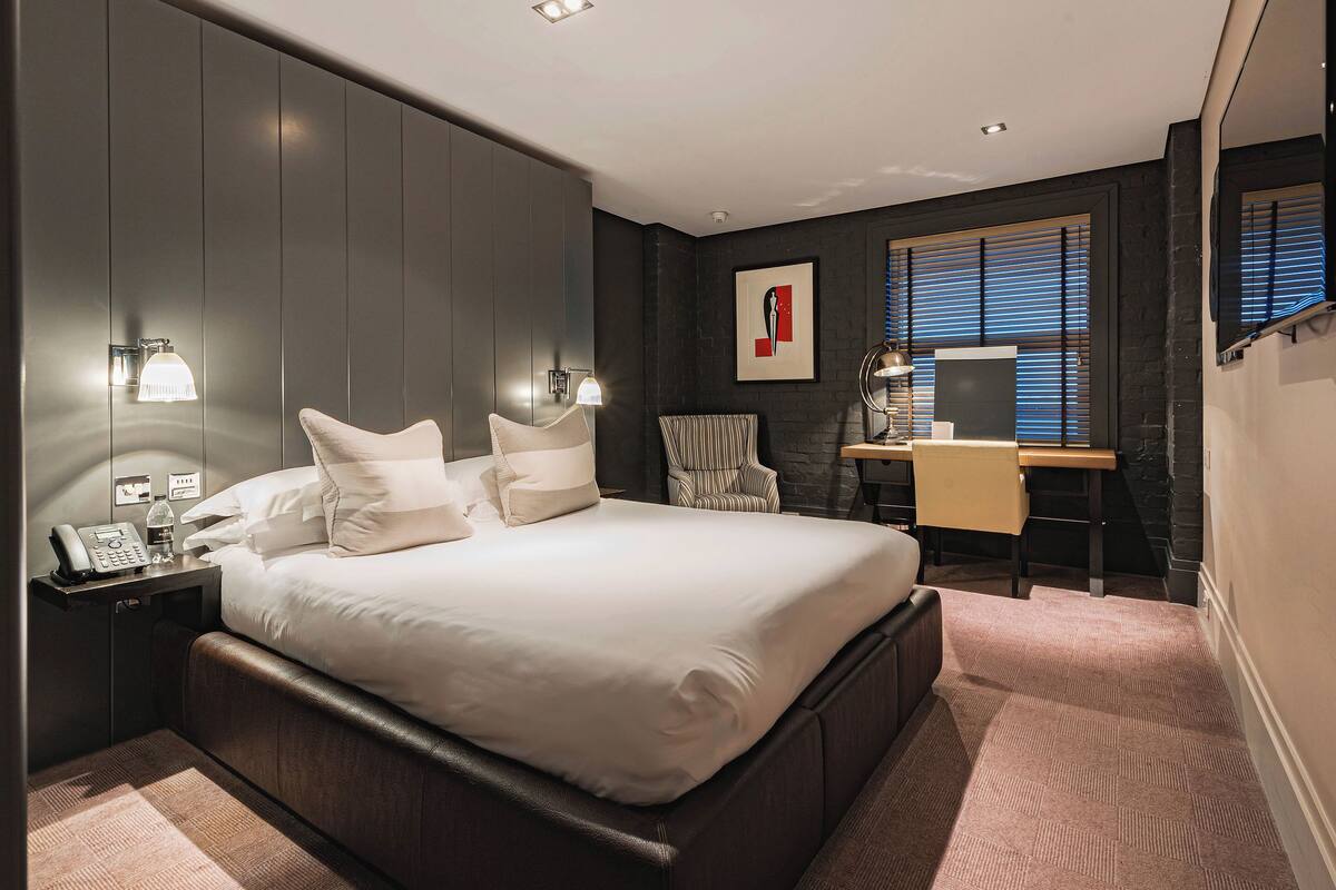 A modern hotel room with a large bed, bedside table with a phone and bottle, a desk with a chair, and a window with blinds.