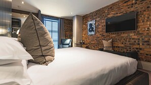 A modern hotel room with a large bed, a flat-screen TV mounted on a brick wall, and a small seating area with a chair and table. - Dakota Eurocentral (Motherwell)