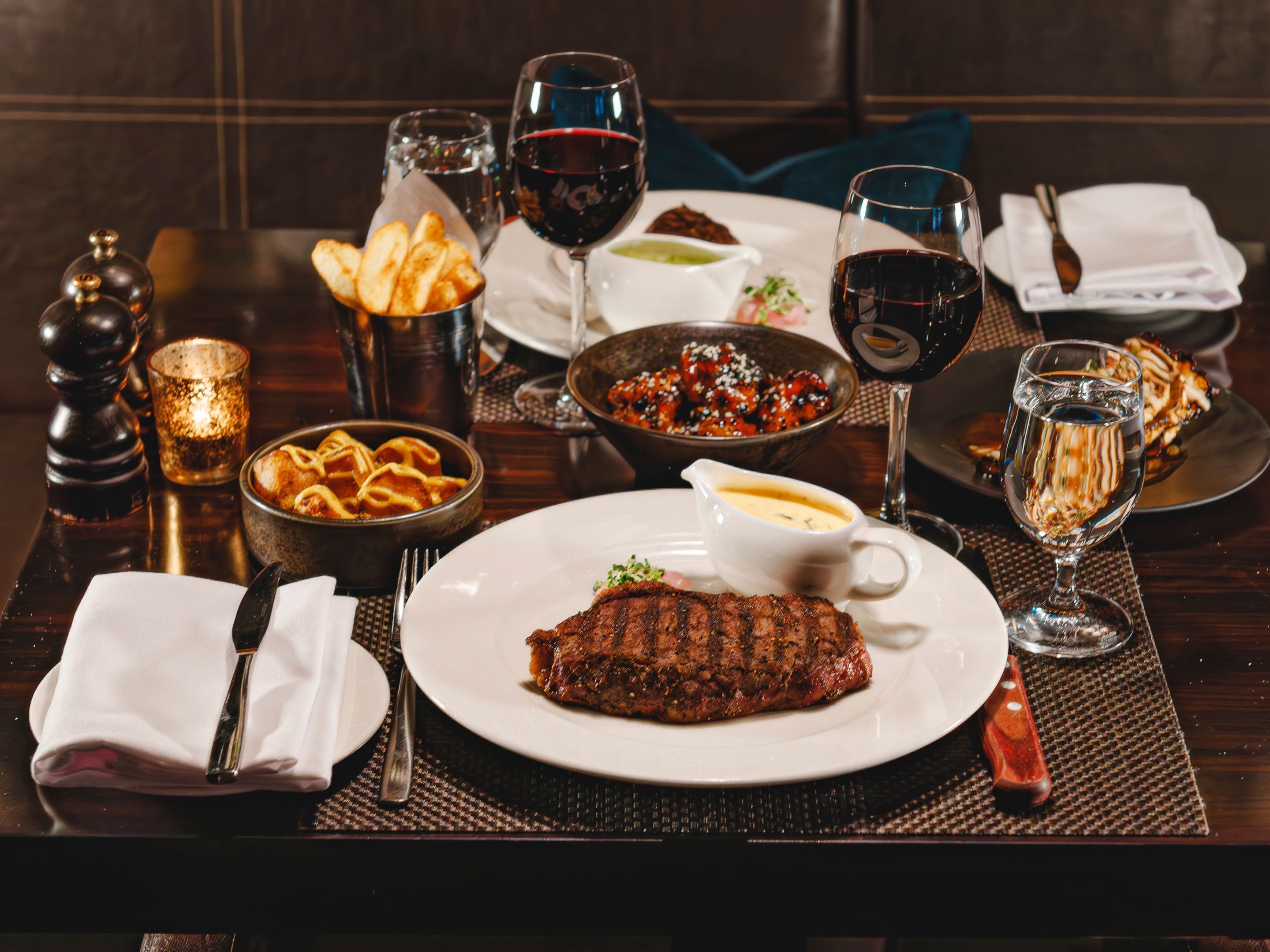 A dining table set with a steak, bread, and various side dishes, along with multiple glasses of red wine.
