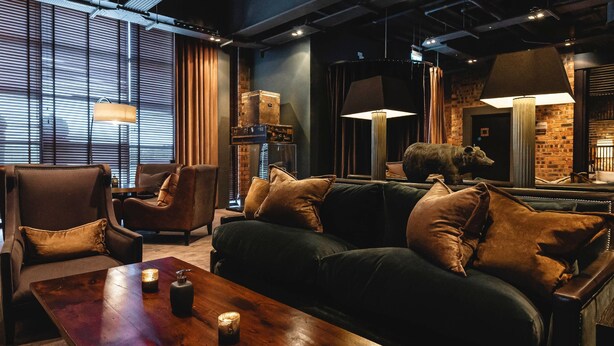 A cozy living room with a dark green sofa, two armchairs, and a wooden coffee table. There are lamps and a brick wall in the background. - Dakota Eurocentral (Motherwell)