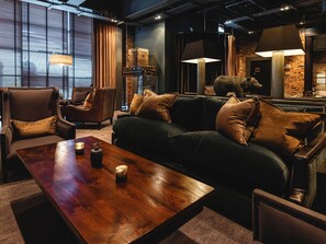 A cozy living room with a dark green sofa, two armchairs, and a wooden coffee table. There are lamps and a brick wall in the background. - Dakota Eurocentral (Motherwell)