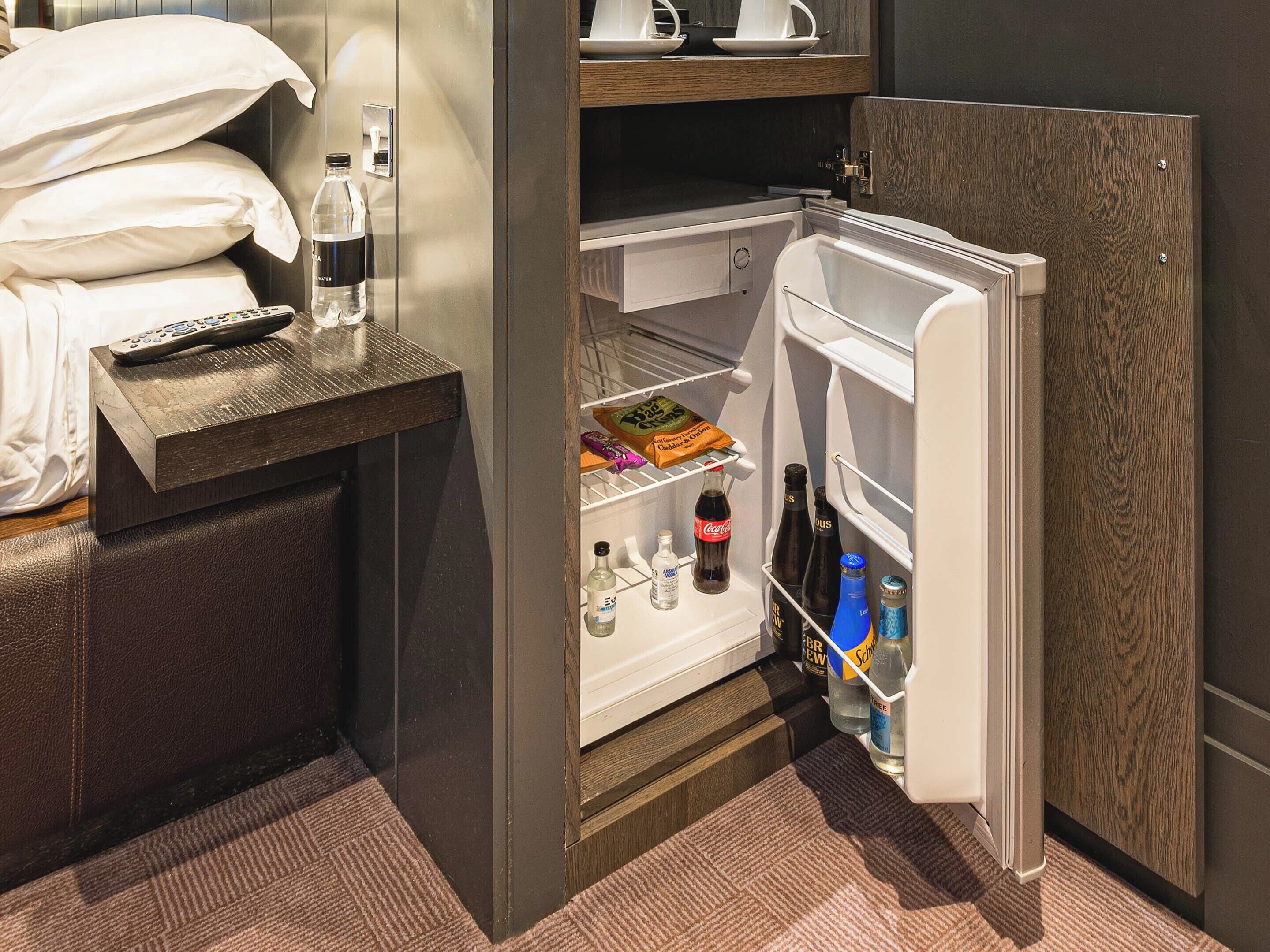 Habitación de hotel con cama, mesita de noche y un refrigerador abierto que contiene bebidas y aperitivos.