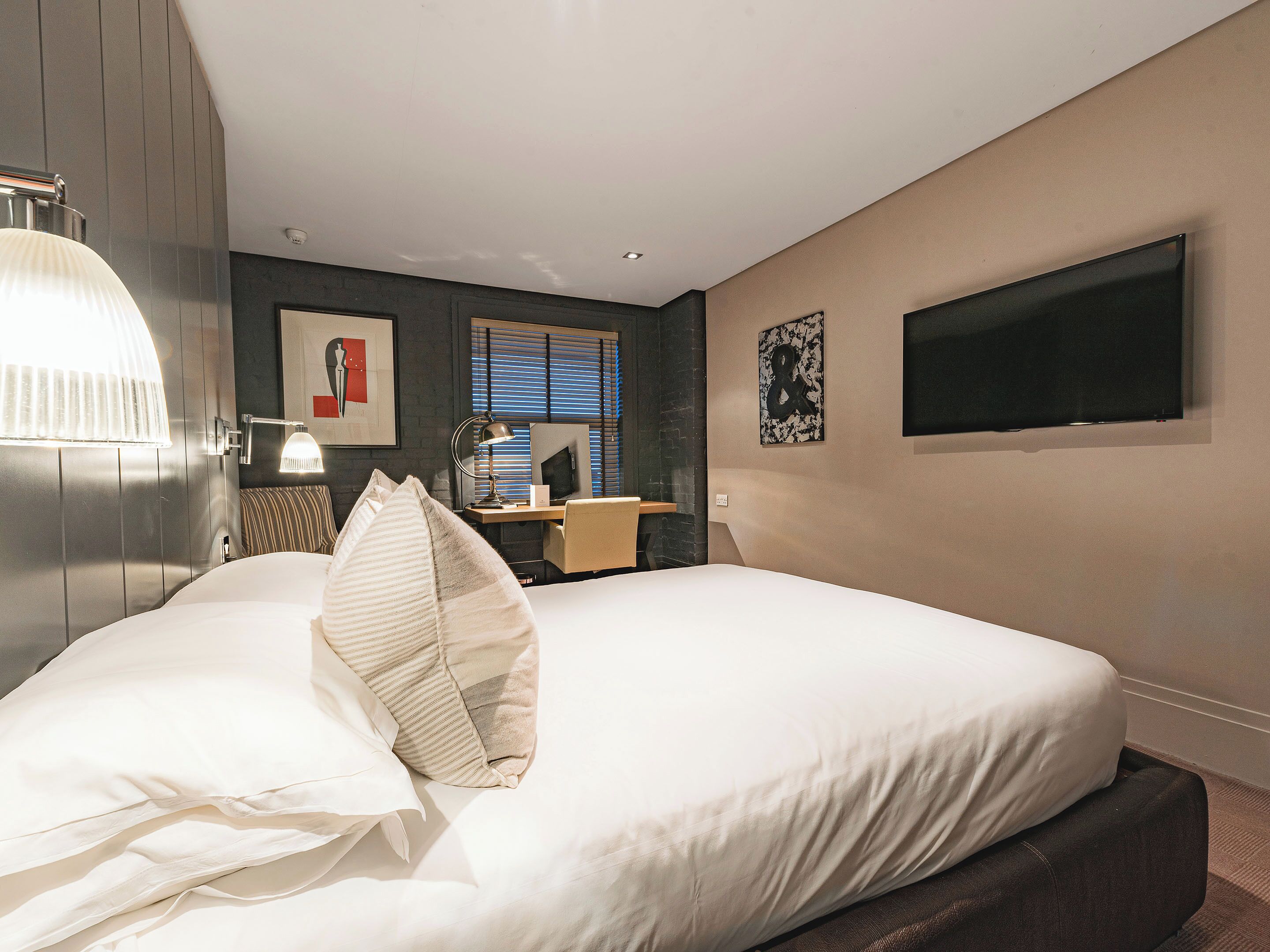 Habitación de hotel con cama, escritorio, silla, lámpara, televisor y obras de arte en las paredes.