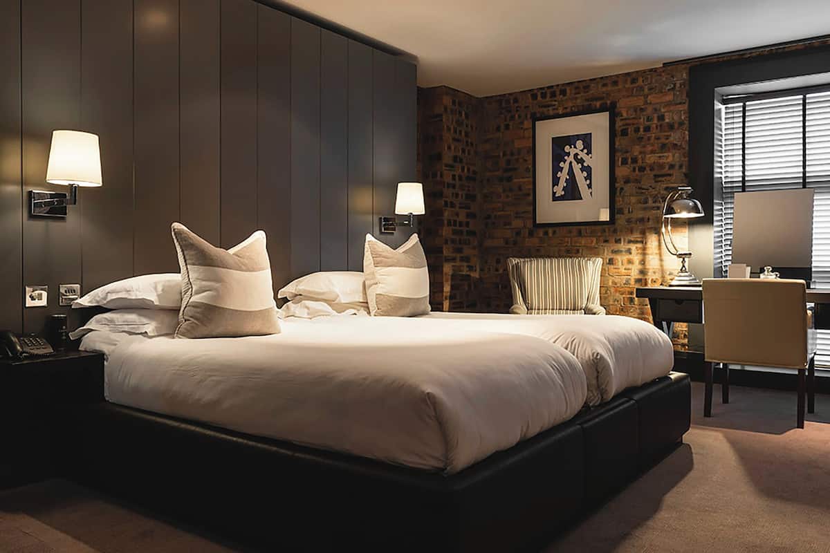 A hotel room with a large bed, two bedside lamps, a desk with a chair, and a brick wall with a framed picture.