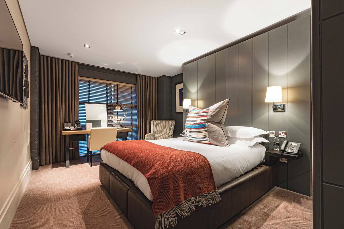 A hotel room with a bed, bedside table, lamp, and a desk with a chair.
