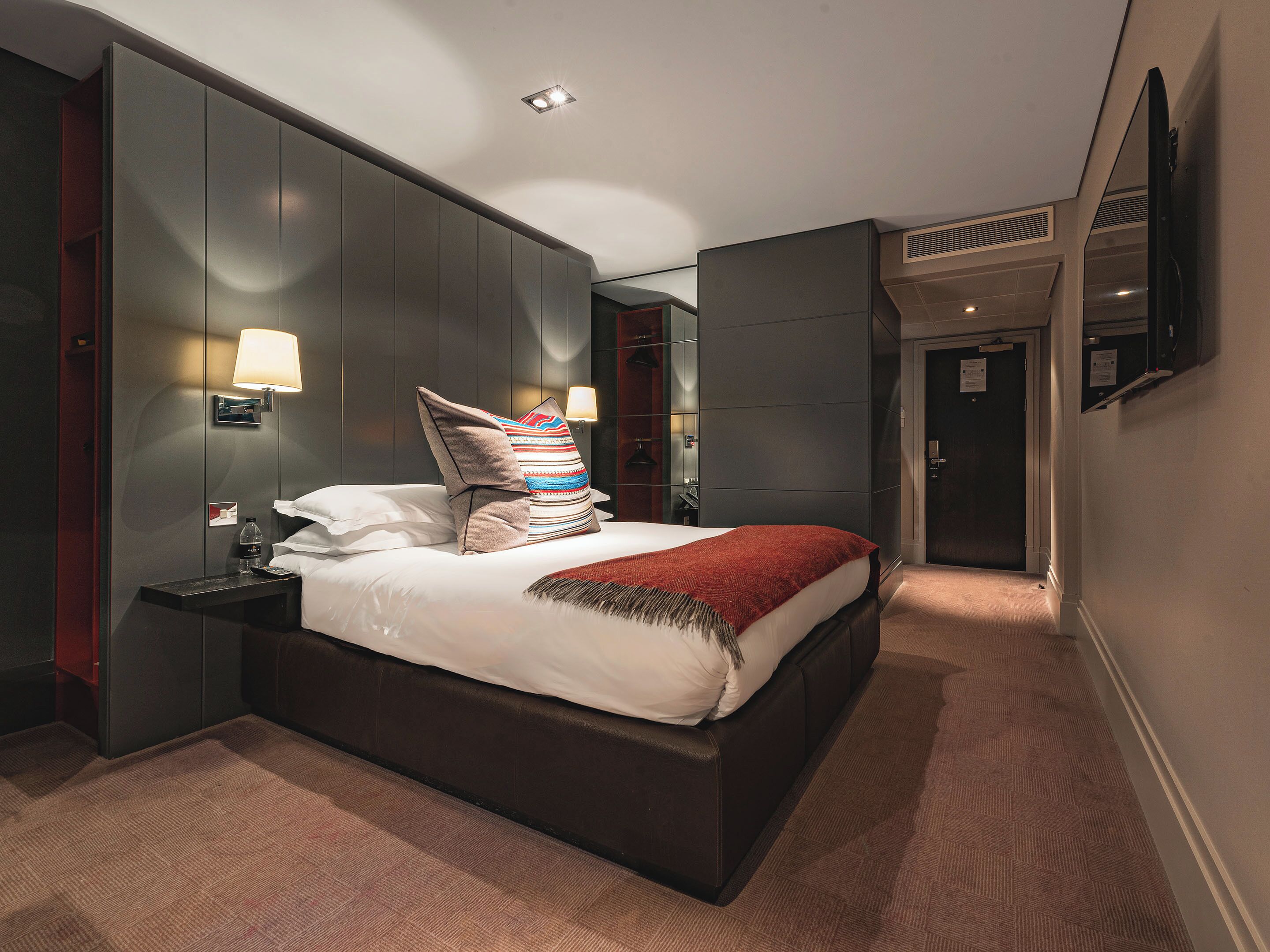 A modern hotel room with a large bed, bedside lamps, a nightstand, and a mirror on the wall.