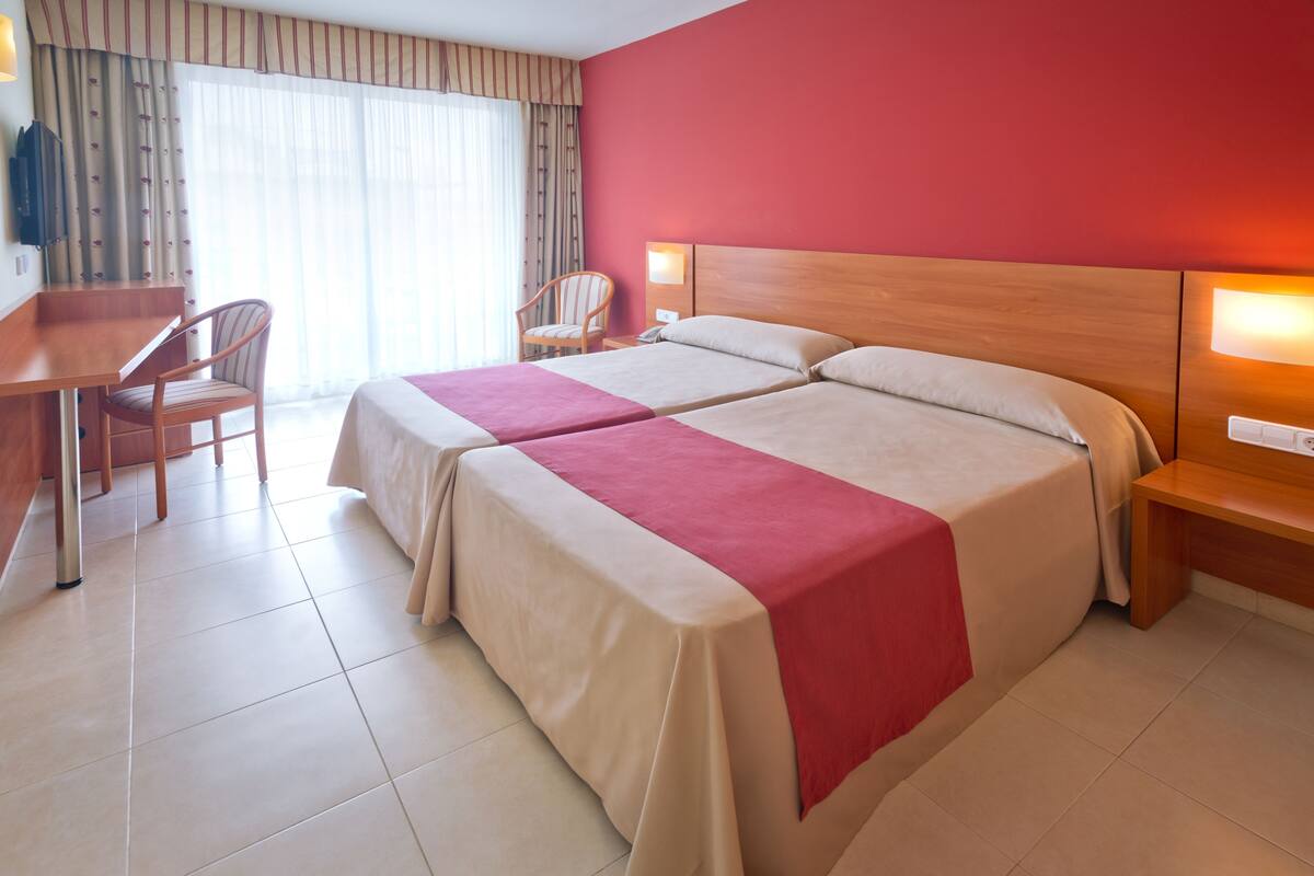 A hotel room with two beds, a desk, a chair, and a large window with curtains.