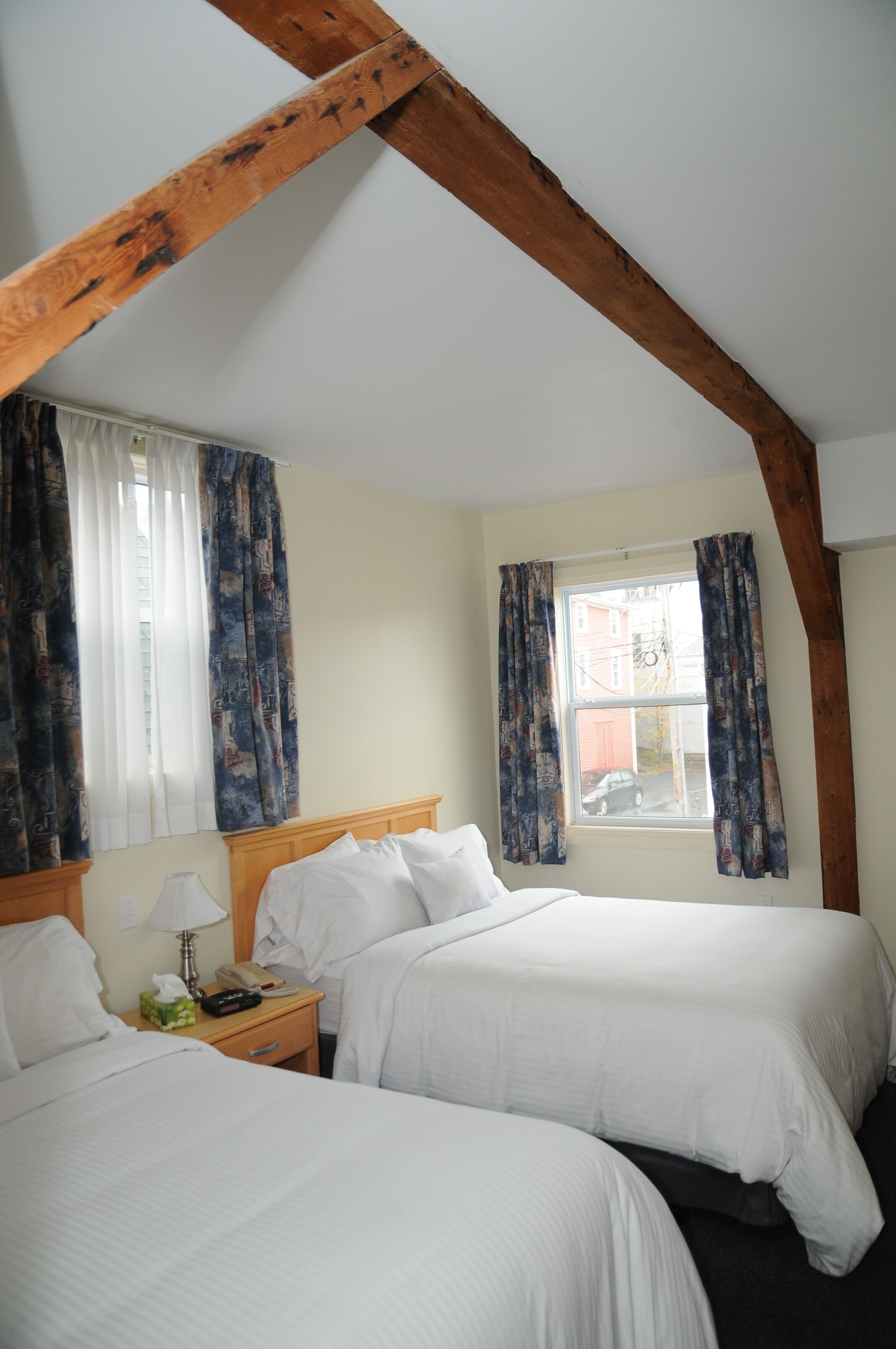 A room with two beds, wooden beams, a window with curtains, a bedside table with a lamp, and a telephone.
