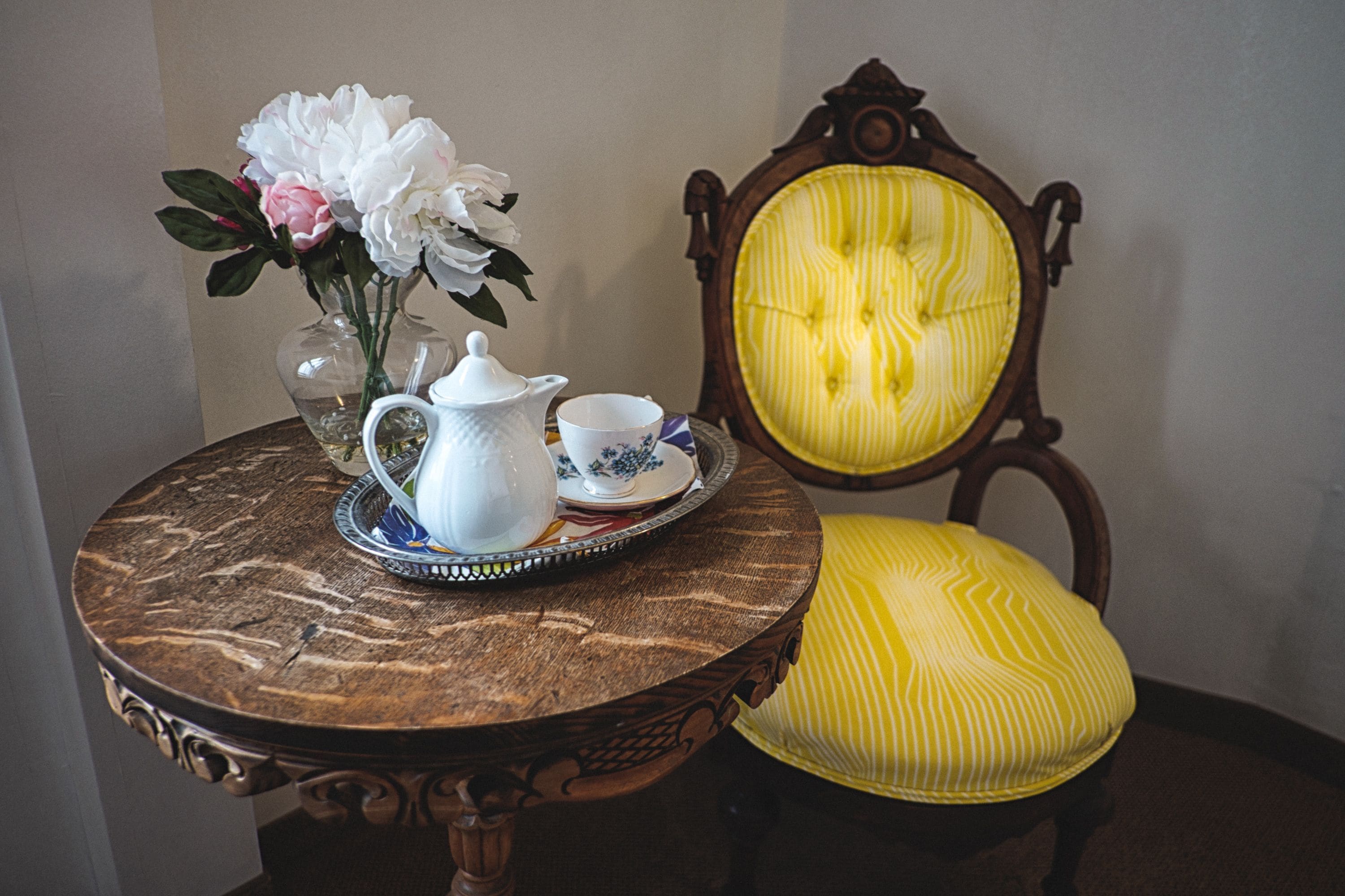A round wooden table with a tray holding a white teapot, a cup, and saucer, and a vase with white and pink flowers. A yellow upholstered chair is next to the table.