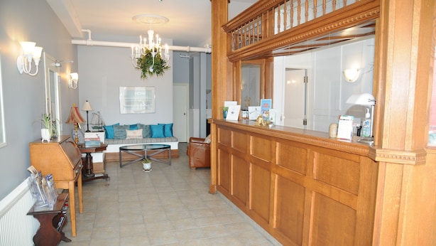 Un espace d’accueil comprenant un comptoir de réception en bois, un lustre, un coin salon avec un canapé et des fauteuils, et un grand miroir au-dessus du comptoir.