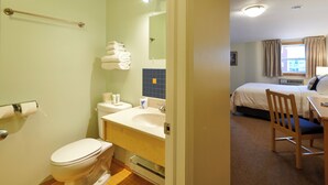 A hotel bathroom with a toilet, sink, and towel rack. The room also includes a bed, a chair, and a desk with a lamp.