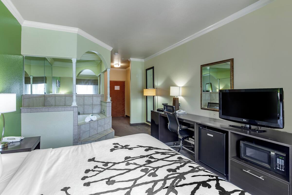 A hotel room with a bed, a desk, a television, a mirror, and a bathroom with a shower and toilet.