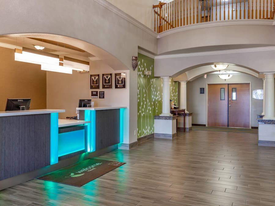 A hotel reception area with a reception desk, a digital display, framed awards on the wall, and a staircase leading to an upper level.